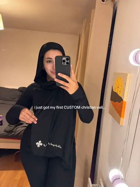 A mirror selfie shows a person in a black hooded outfit holding a black veil adorned with text and a small design. The person is smiling, holding a phone in their other hand taking the picture.