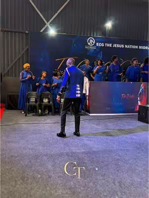A stage with a male figure facing away from the camera. Behind the figure are several people standing in blue outfits and towards the wall there is sign with ECG. The floor is covered with a gray carpet. The image is well-lit with a focus on the stage area.