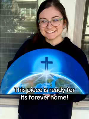 A woman with glasses smiles, holding a canvas print depicting a cross set against a blue, semi-circular background of the Earth and outer space, with text overlayed
