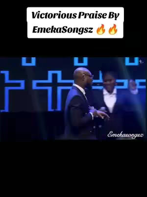The image features a stage with two people in focus. Text overlays at the top read "Victorious Praise By EmekaSongs" in a white box. Below, in the background, a stylized blue cross design sits behind the main subjects. One man, dressed in a suit, is in the foreground, and a woman, also in a suit, stands beside him. The image seems to be taken during a concert or church service with the artist.