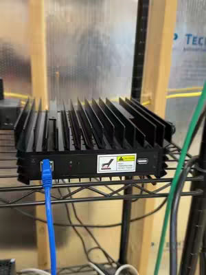 The image shows a black electronic device with a series of parallel, slotted ridges across its top, suggesting a heat sink design. It's positioned horizontally in the frame, with various cables, including a blue Ethernet cable, connected to its front side. The setup is on a black wire shelf. Behind the device, a wooden frame and a semi-transparent, possibly plastic, backdrop are visible, and there are visible stickers on the device. Overall, the image conveys a sense of a technological or electronic setup.