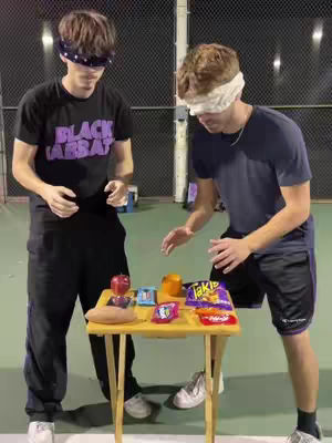 Two young men, both wearing blindfolds, stand on a tennis court. They are facing each other with a small table between them, laden with snacks. The man on the left wears a black t-shirt with a band logo and black pants. The man on the right is in a navy shirt and black shorts. The background includes a mesh fence.