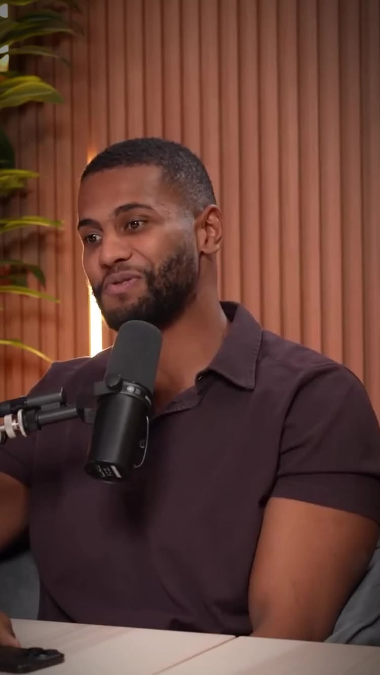 The image shows a man in a casual setting, likely a studio or a room with wooden paneling as a backdrop. He is wearing a dark purple shirt and is positioned in front of a microphone, suggesting he is either speaking or recording a podcast. His facial expression is neutral, with a slight hint of a smile, and he has facial hair. The lighting appears to be soft and diffused, creating a balanced shot.