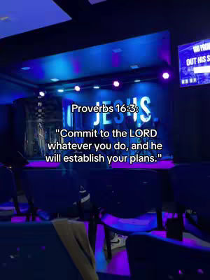 The image shows a stage with a large 'JESUS' sign, lit by various stage lights. Text overlays include a Bible verse. The composition is dark, with an emphasis on the illuminated stage and text.