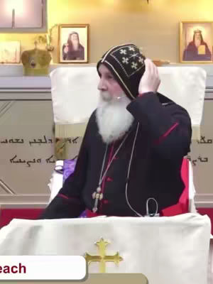 A man in religious attire is seated, adjusting his head covering. He has a long white beard and is wearing a dark robe with red accents. The background includes framed images and a decorative setting.