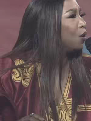 A woman with long dark hair and makeup is seen from the side, singing into a microphone. She is wearing a maroon outfit with gold embroidery on the shoulders. The background is a solid, muted red.