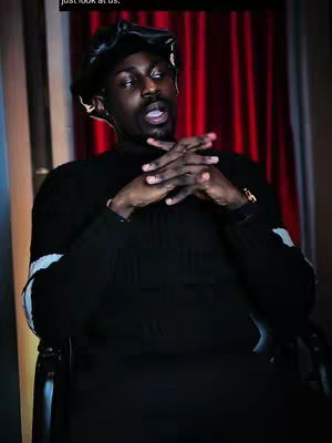 The image features a Black man seated in a chair, positioned against a backdrop of a red curtain. The man is wearing a black fitted cap, a black turtleneck sweater, and a gold watch. He has his hands clasped together in front of his face with a serious expression.