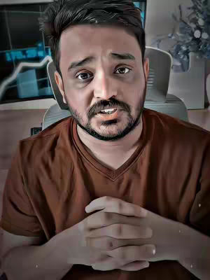 A man with a beard and brown shirt sits facing the camera, with hands clasped and a serious expression. A computer screen is behind him, displaying charts.