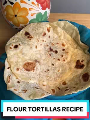 The cover image showcases a stack of flour tortillas in a teal-colored bowl. The tortillas are light in color with visible brown spots and slight imperfections. In the background, part of a decorative, colorful ceramic jar is visible, placed on a wooden surface. Below the tortillas, a blue rectangular banner displays the text "FLOUR TORTILLAS RECIPE" in white.