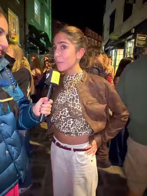 A woman is being interviewed on the street at night. She's wearing a brown leather jacket, a patterned top, and white pants. The background is blurry with other people and buildings.