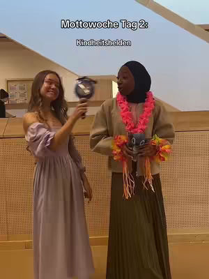 The cover image shows two young women in costumes for a theme day. One, on the left, is dressed in a lilac dress and smiles, holding up a round object resembling a fan. The other, on the right, is wearing a dark hijab, a beige jacket, and a long green skirt. She has a pink floral lei around her neck and is holding a stuffed toy in her hands. The background is a light-colored room with a slightly angled ceiling and wall paneling.