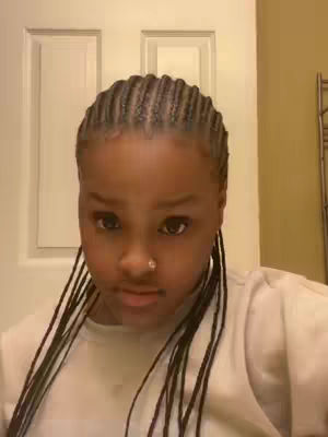 A close-up selfie features a young person with neatly styled braids. The background is an out-of-focus interior, likely a room or bathroom. The person has a small nose ring and is looking directly at the camera with a neutral expression, the upper body is visible, and the person is wearing a white top.