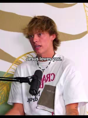 A young man with light brown hair is the focus, his face centered. He's wearing a white t-shirt with a graphic. A microphone is positioned in front of him. Text overlay reads, 'Jesus loves you.' The background is a blurry wall.