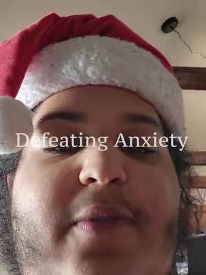 A close-up selfie shows a person wearing a red Santa hat with a white trim. The person has a light complexion, dark hair, and a bit of facial hair. The text 'Defeating Anxiety' is overlaid on the image. The background is slightly out of focus. The lighting seems to be indoor and natural.