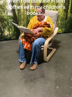 A woman sits in a light-colored wooden chair, reading a book. She is wearing a yellow sweater, blue jeans, and tan shoes. A blanket with an orange pattern is draped over her lap, and behind her is a backdrop of a forest scene. The overall composition is warm and inviting.