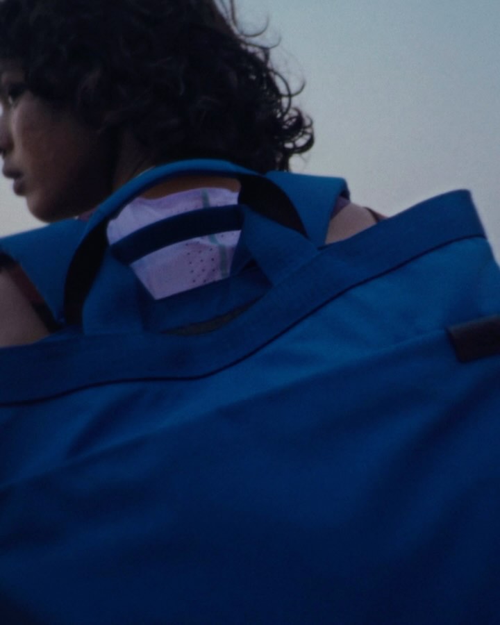 The image shows a person, seen from the back, wearing a blue tote bag. The person is wearing a red shirt; the top of the person's head is visible. The bag has a gray interior, and its left strap rests over the person's left shoulder. A white label sits beneath the the bag's strap. The scene seems to be outdoors, possibly at dusk, indicated by the muted colors and sky.