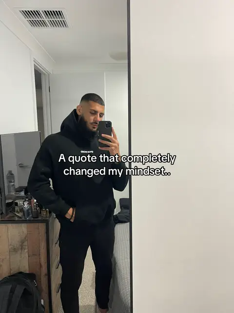 A man in a black hoodie and pants is holding a phone in front of a mirror, taking a selfie. The background is a simple modern room with a white wall, a large mirror reflecting the man, and a door on the left side. There’s a text overlay on the image.