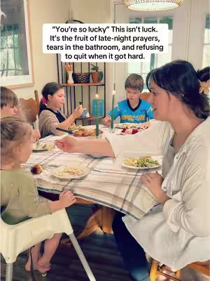 A well-lit photo captures a heartwarming family scene around a dining table. A woman, appearing to be the mother, is the central figure, partially visible from the side as she interacts with the members. Surrounding her are several children, engaged in their meal. The table is set with plates of food and candles, suggesting a special occasion. The background is a mix of a window and decorative elements, contributing to the inviting atmosphere.