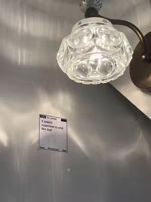 The image shows a portion of a wall with a light fixture and a sticky note. The light has a clear, multi-faceted glass shade and is positioned near the top of the frame. The sticky note is rectangular and contains text, situated below the light fixture. The wall is a muted gray color. The composition is vertical, with the light and the sticky note occupying the majority of the space.