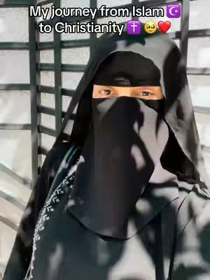 A person wearing a black hijab and niqab is the central focus. The individual's eyes are visible, peering out from the dark fabric. The background features a partial view of a metal gate or lattice.