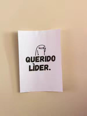 A white piece of paper is affixed to a beige wall. On the paper, the words "QUERIDO LÍDER." are written in black, bold text. Above these words is a crudely drawn cartoon figure. The lighting is soft, and the paper casts a slight shadow against the wall.