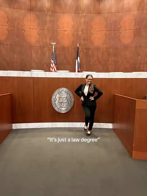 A woman in a black suit stands in a courtroom-like setting, smiling at the camera. The background features a wooden wall and a seal.