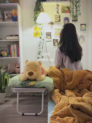 The image showcases a bedroom scene with a young woman facing away from the camera, sitting on the edge of a bed covered with a brown blanket. A shelf with books is visible on the left side, and a Winnie the Pooh plush toy lies on a stool in the foreground. Additional decor includes pictures on the wall and a floor lamp. The overall composition depicts a cozy and relaxed atmosphere.