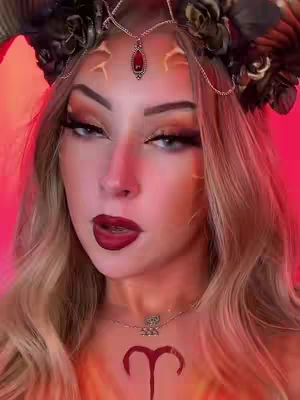A close-up shot of a woman's face, adorned with elaborate Aries-themed makeup. She sports a dark maroon lipstick, heavy eye makeup with a prominent wing, and a decorative headpiece featuring horns and floral elements. Her skin is highlighted, and an Aries symbol is painted on her chest. The background is a gradient of reddish-pink.