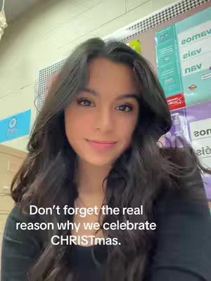 A woman with long dark hair looks at the camera with a slight smile. Text below reads, 'Don't forget the real reason why we celebrate CHRISTmas.' The background is a mix of office decor, including what appears to be calendar or posters.