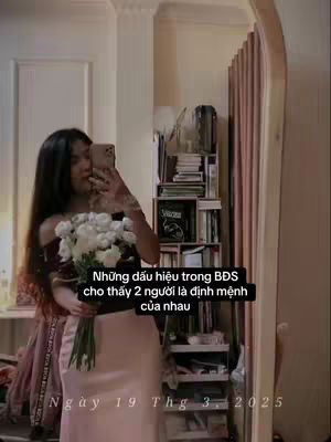 A woman is shown in a room, appearing in a mirror reflection. She is holding a bouquet of white flowers and is taking a selfie with her phone. Text is overlaid, and also a date is shown at the bottom.