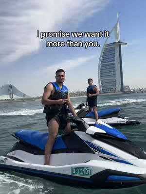 Two men are on jet skis in the ocean with a cityscape and large building in the background.