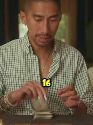 A man is seated at a table, shuffling a deck of cards. He is wearing a light button-down shirt with a green and white checkered pattern and a thin gold chain. Overlayed on the image is a yellow number '16.' The background appears to be a room with wooden trim.