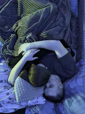 A top-down shot shows a couple embraced on a bed, covered by a textured gray blanket. The individuals are close together, with one's head resting on a patterned pillow. The lighting is soft, with cool tones dominating. The overall impression is intimate and cozy.