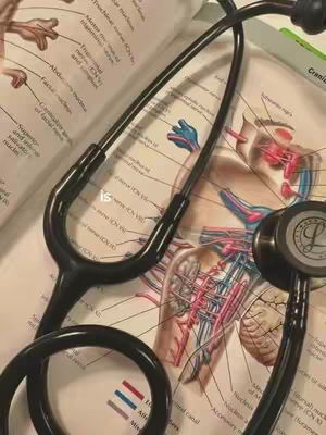 A close-up shot features a black stethoscope draped across an open medical textbook. The book displays detailed anatomical illustrations in a mix of colors and text. The stethoscope's earpieces and chest piece are visible, creating a foreground element that contrasts with the book's busy background. The lighting appears natural, highlighting the textures and colors of both the medical tool and the educational material. The overall composition is intended to show a medical environment, and it could target medical students.