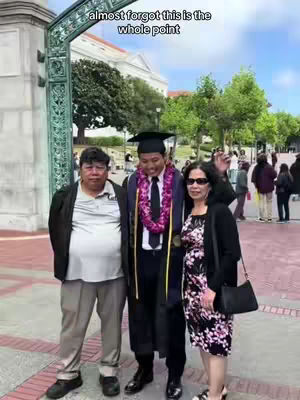 A graduate, wearing a cap and gown, stands between his parents. The graduate is smiling, and his parents are also smiling. The background shows a building, a green archway, and people walking around.