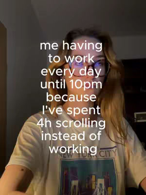 A woman with long, light brown hair and glasses is in a selfie-style video. Overlaid on the image is white text conveying a relatable message about work and scrolling on the phone. She is wearing a graphic t-shirt, and the background is dimly lit.