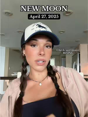 A woman with dark hair in pigtails wearing a baseball cap is centered in the frame. The cap has a horse logo on the front. Overlay text at the top reads 'NEW MOON' and 'April 27, 2025.' Additional text is in the corner 'Check sun, moon. RISING'. The background is a softly lit interior with light fixtures visible.