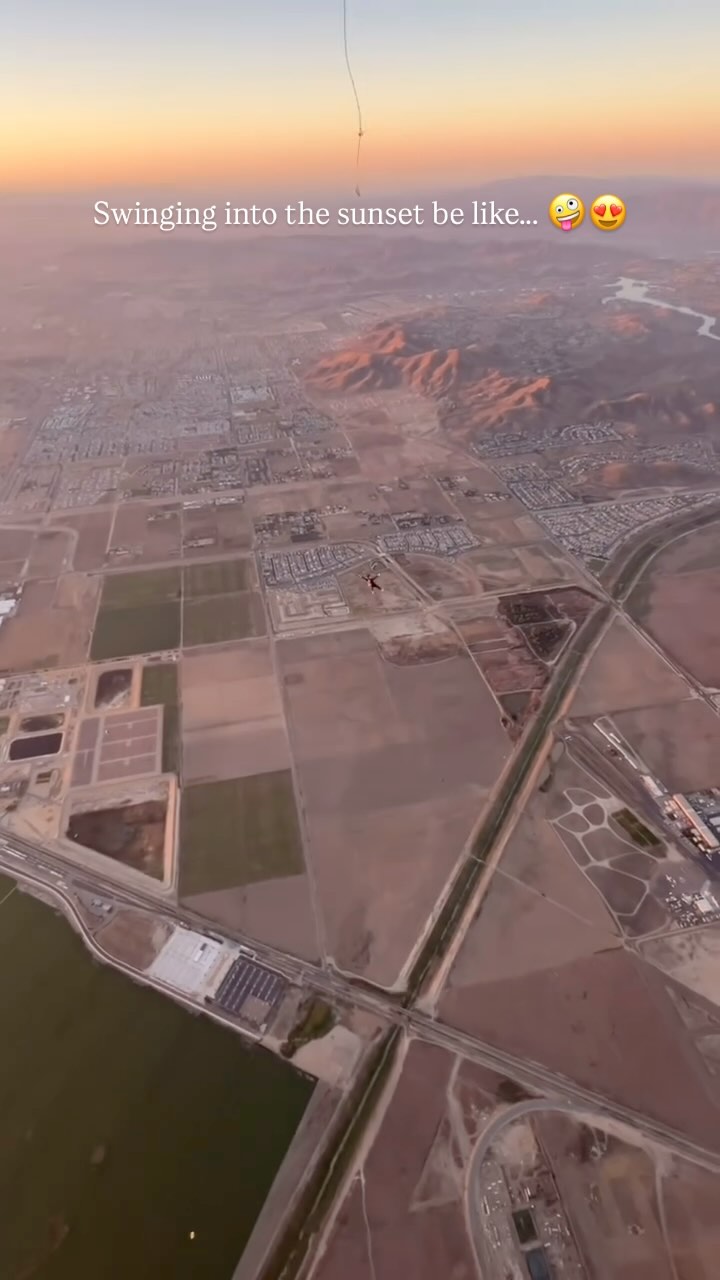 Aerial view of a city and agricultural landscape during sunset. The sky transitions from orange to a lighter hue, with a skydiver visible. The cityscape and fields are detailed with roads and buildings, all tinged with warm light.