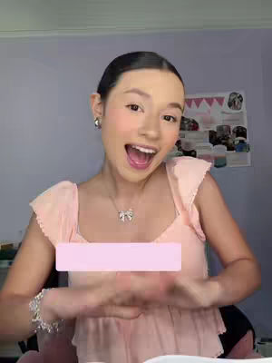 A woman with dark hair, wearing a light pink top and a necklace with a bow pendant, is looking at the camera with an open mouth and hands clasped together. Her expression is excited. A pink rectangle covers part of the image's foreground. The background contains a shelf with items and a light purple colored wall.