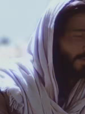 A close-up shot of a man, presumably Jesus, with light skin and a beard. He is wearing a white hood, and most of his face is in shadow, with only a portion of his cheek and beard visible. The lighting is soft, suggesting a serene or spiritual setting.