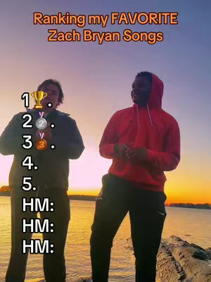 Two young men stand side-by-side, positioned on a rocky outcrop with an ocean backdrop as the sun sets, giving a warm, orange glow to the sky and water. The man on the left is holding a trophy and medal icons, and is wearing a dark jacket. The man on the right is wearing a red hoodie and black pants. Text overlaid at the top of the image says "Ranking my FAVORITE Zach Bryan Songs." Vertical text along the left side reads "1, 2, 3, 4, 5" followed by "HM: HM: HM:"