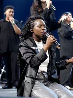 The cover image features a woman of color with long curly hair and a black blazer, holding a microphone and singing on a stage with a worship team in the background. The setting appears to be a church service with a spotlight. The composition highlights the woman as the central figure, emphasizing the emotional connection to the song. The overall look is one of spiritual expression.
