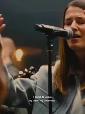 A woman with long brown hair, wearing a light-colored jacket, singing into a microphone. She has her eyes closed and her hands are raised in a gesture of worship. Behind her, another person and a soft light are blurred.