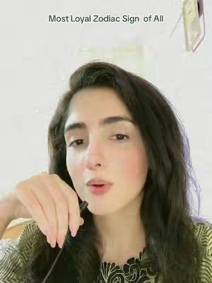 A woman with long dark hair is in the center of the frame, angled slightly towards the camera. The image is brightly lit, and the woman is looking towards the camera with a slight expression. A thin black microphone is held near her mouth. The background is neutral, with a bright, slightly blurred effect.