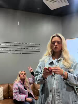 Two young women are present in the cover image. The main focus is on the woman to the right, who is holding something small, possibly food or a cosmetic item. She is wearing a denim jacket over a blue top and various bracelets. A second woman, wearing a quilted patterned jacket, makes a peace sign in the background. They appear to be indoors, and there is text on the wall and some brown furniture in the background.