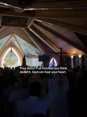 A dimly lit church interior with a high, arched ceiling and stained-glass windows. Silhouetted figures of people are in the foreground, focusing on a dark cross.