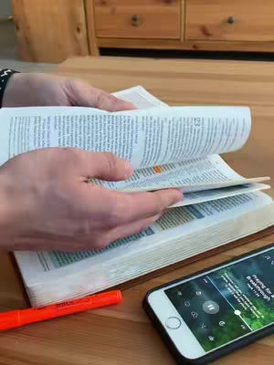 A person's hands are shown holding a Bible and flipping the pages. There is a smartphone with app interface showing on the table, and a bright orange marker next to it.