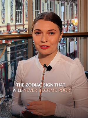A woman with fair skin and brown hair is centered in the frame, looking directly at the camera. She is wearing a white collared shirt. Below her, the text overlay reads, 'THE ZODIAC SIGN THAT WILL NEVER BECOME POOR.' The background includes a blurred interior setting with windows and decorative elements.