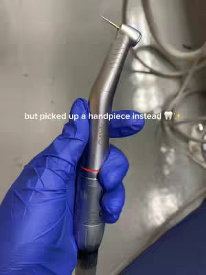 The image shows a hand wearing a blue medical glove holding a silver dental handpiece. The background is a stainless steel surface, possibly in a dentist's office. Text overlayed on the image reads: "but picked up a handpiece instead".