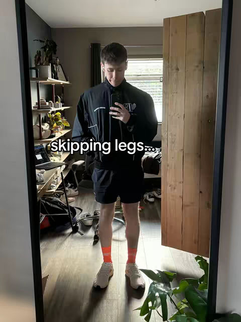 A man stands in a room, reflected in a large mirror. He is wearing a black jacket, black shorts, and orange socks. The room appears to be a bedroom or home office, with a window visible in the background. White text overlay 'skipping legs'.